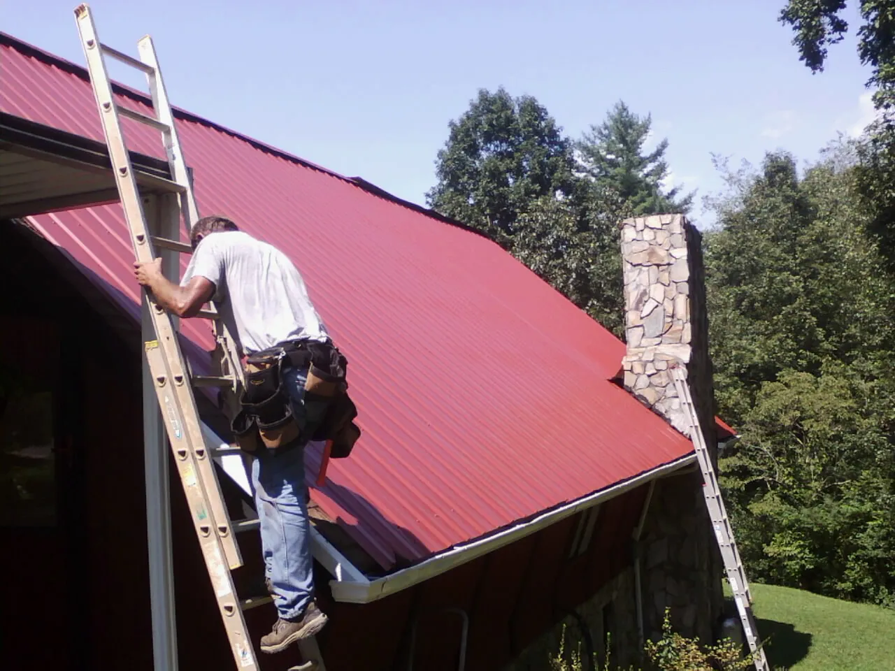 Roofer on a ladder beside a red metal roof and stone chimney — Soffit & Fascia Repair by Roofing Brothers in Carrsville